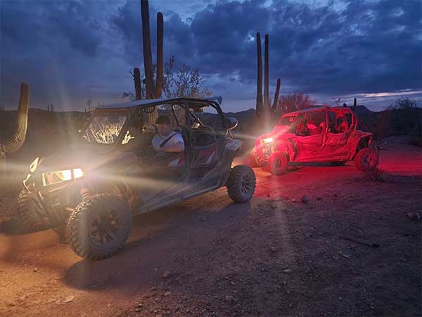Two side-by-side UTVs ride through the desert at sunset with their lights on, illuminating a dirt road